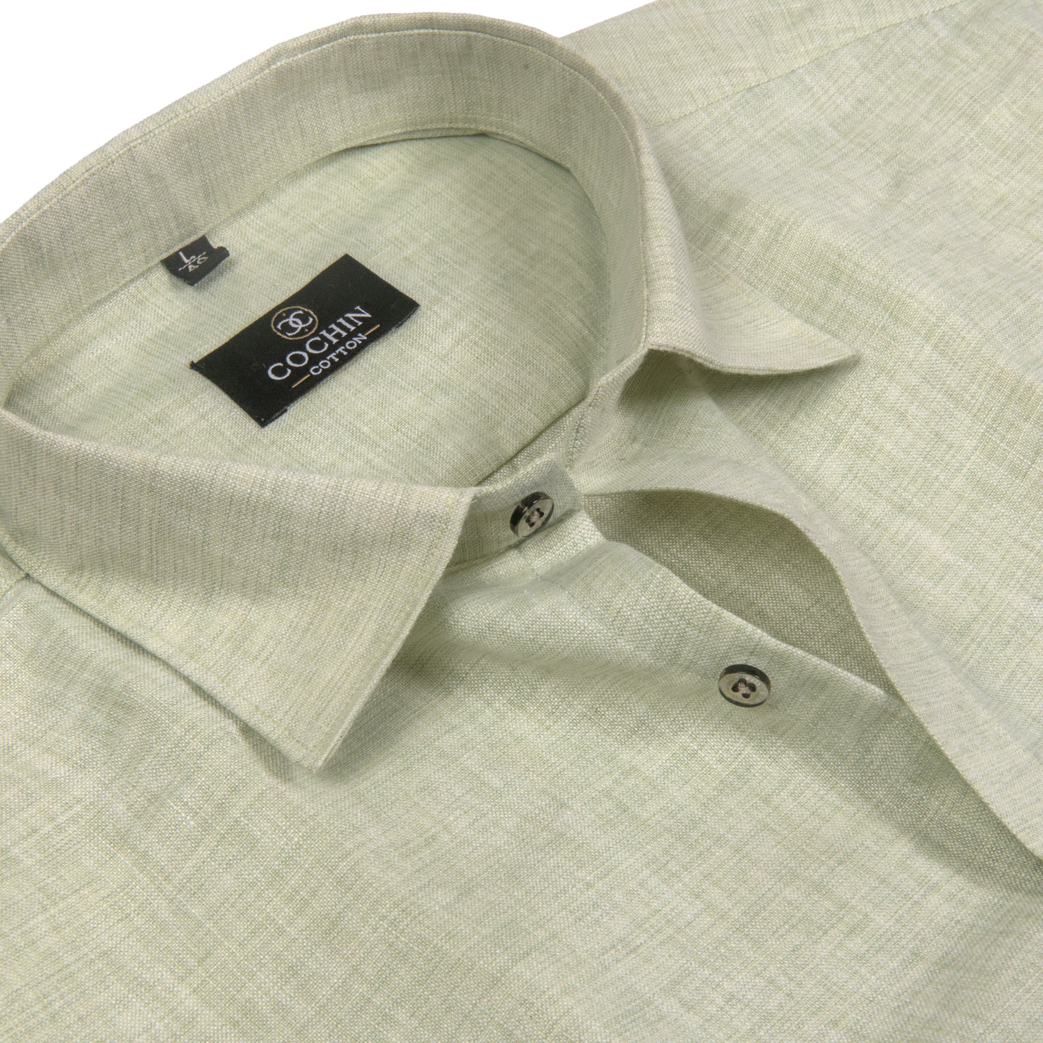 A close-up of a light sage green linen shirt by Cochin Couture, showing the textured fabric, collar, and black brand label.
