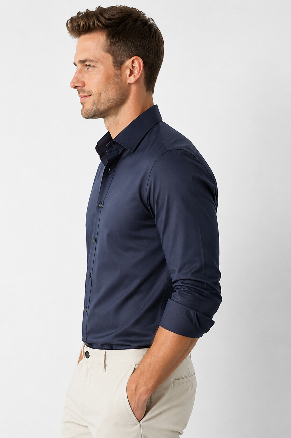 A man in side profile wearing a dark navy blue long-sleeve button-down shirt and light beige trousers, photographed against a light gray studio background.