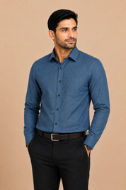 A man wearing a long-sleeve blue button-down shirt with black trousers, standing against a solid beige background.