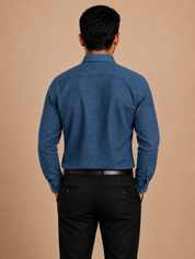 A man viewed from behind wearing a blue long-sleeve button-up shirt and black trousers, standing against a solid beige background.