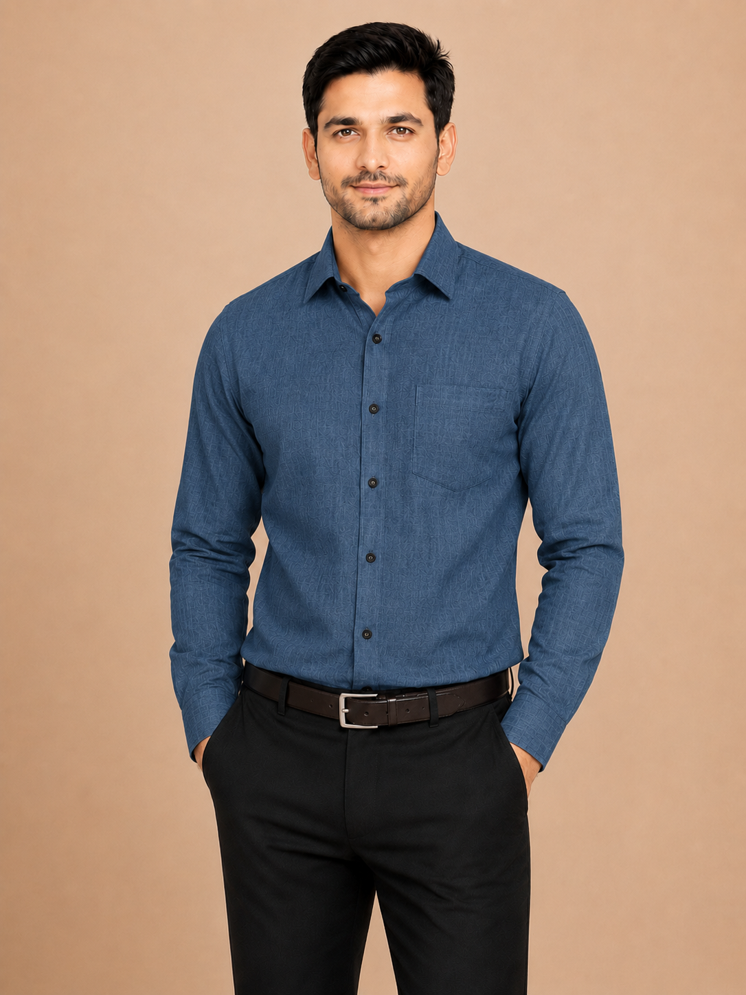 A man wearing a long-sleeve blue textured button-down shirt with black trousers and a brown belt, standing against a solid beige background.