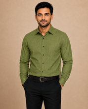A man wearing a long-sleeve olive green button-down shirt and black trousers, standing against a solid beige studio background.