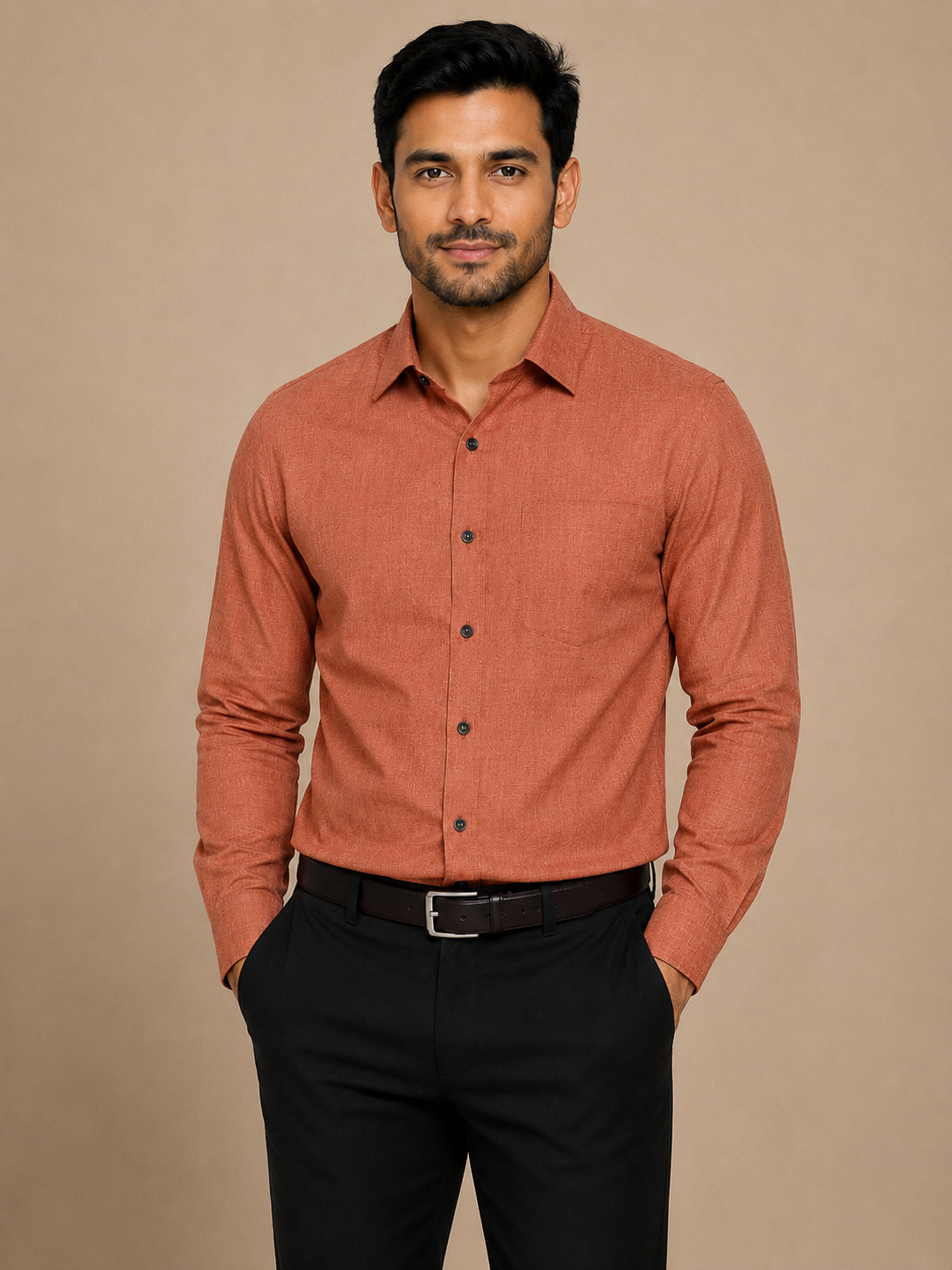 A man wearing a long-sleeve button-down shirt in a muted terracotta or burnt orange color, paired with black trousers and a black belt, standing against a solid beige studio background.