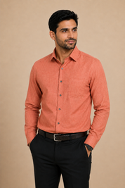 A man wearing a long-sleeve, button-down linen shirt in a muted coral-orange color, paired with black trousers and a black belt, standing against a solid beige studio background.