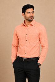 A man wearing a peach-colored long-sleeve button-down shirt and black trousers, standing against a solid beige background.