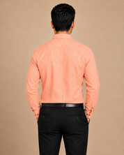 A man viewed from behind wearing a peach-colored long-sleeve textured shirt and black trousers, standing against a solid beige background.