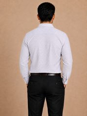 A man viewed from behind wearing a light gray and white micro-patterned long-sleeve button-down shirt, paired with black trousers and a dark brown belt, against a solid beige background.